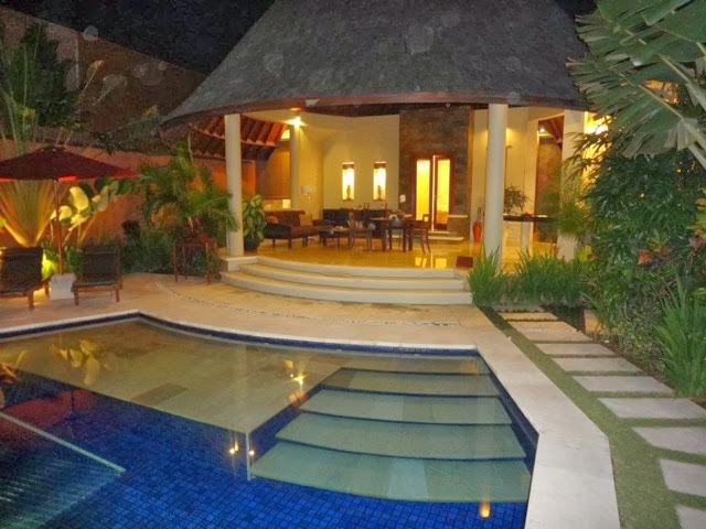 Illuminated outdoor pool at night beside an open-sided pavilion with tropical plants, warm lighting, calm mood