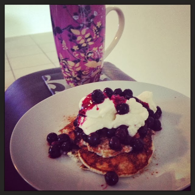 Plate of pancakes topped with cream and blueberries next to a floral patterned mug on a table, warm cozy indoor tones